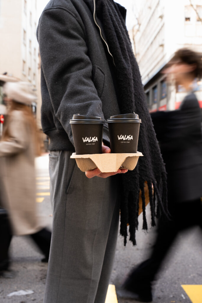 Persona sosteniendo dos cafés para llevar en vasos Valka Coffee Stop en portavasos, escena urbana en la calle.