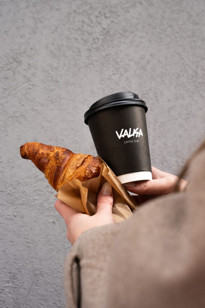 Manos sosteniendo un croissant y un café para llevar en vaso Valka Coffee Stop, desayuno para llevar.
