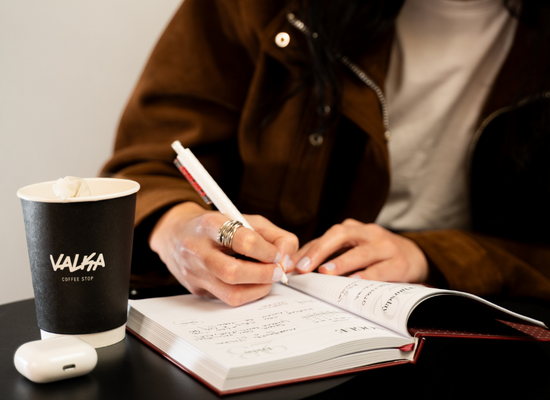 Persona escribiendo en una libreta con un café para llevar en vaso Valka Coffee Stop sobre la mesa, momento de trabajo en cafetería.